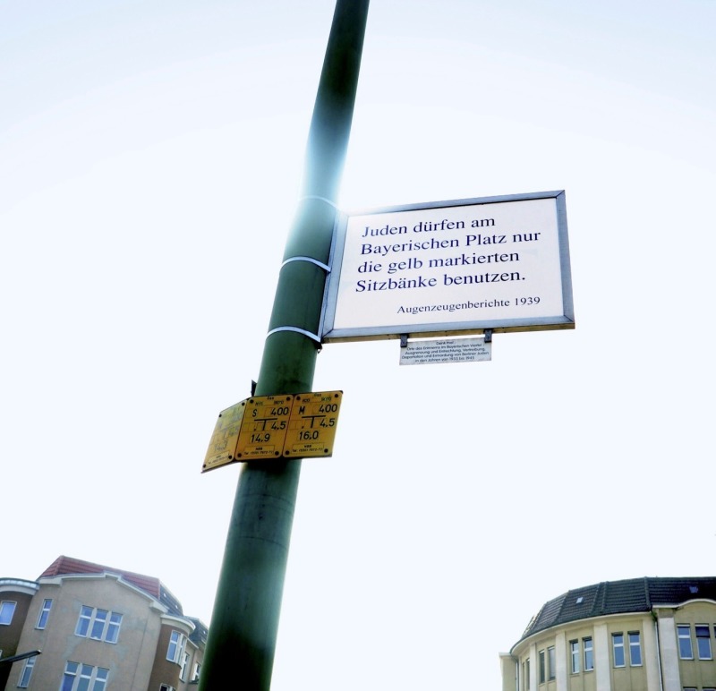 Auf der anderen Seite des Schildes ist zu lesen: „Juden dürfen am Bayerischen Platz nur die gelb markierten Sitzbänke benutzen.“