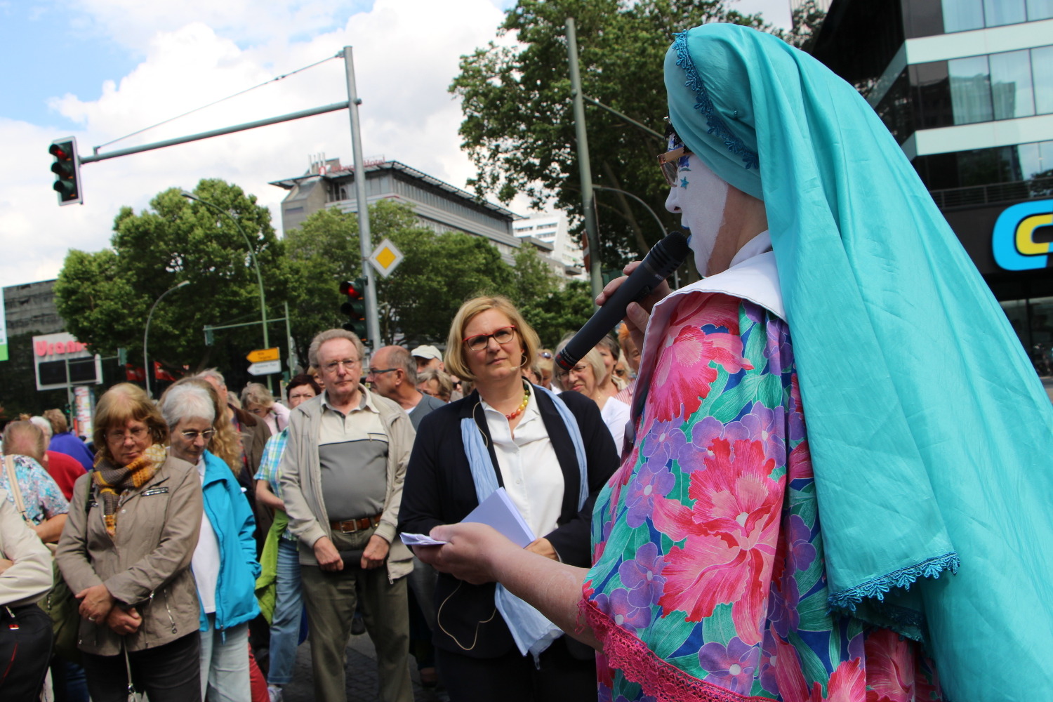 Buntgekleidete Frau mit türkisem Kopftuch und weißbemaltem Gesicht spricht vor Menschen.