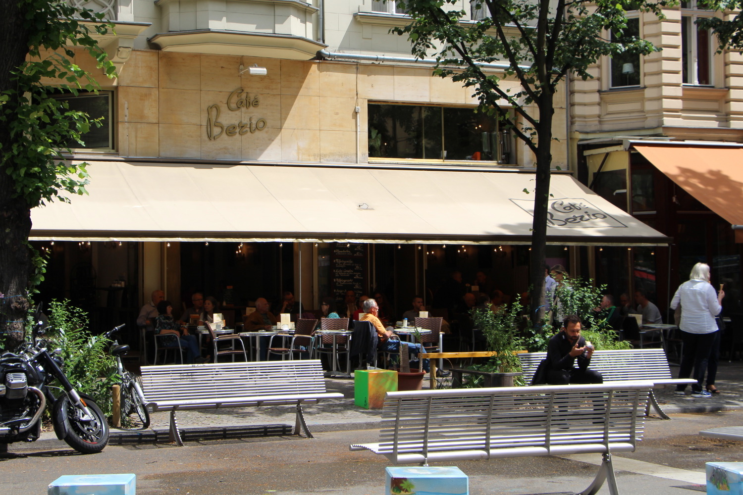 Café mit Tischen und Stühlen draußen und davor ein breiter Gehweg mit Bänken.