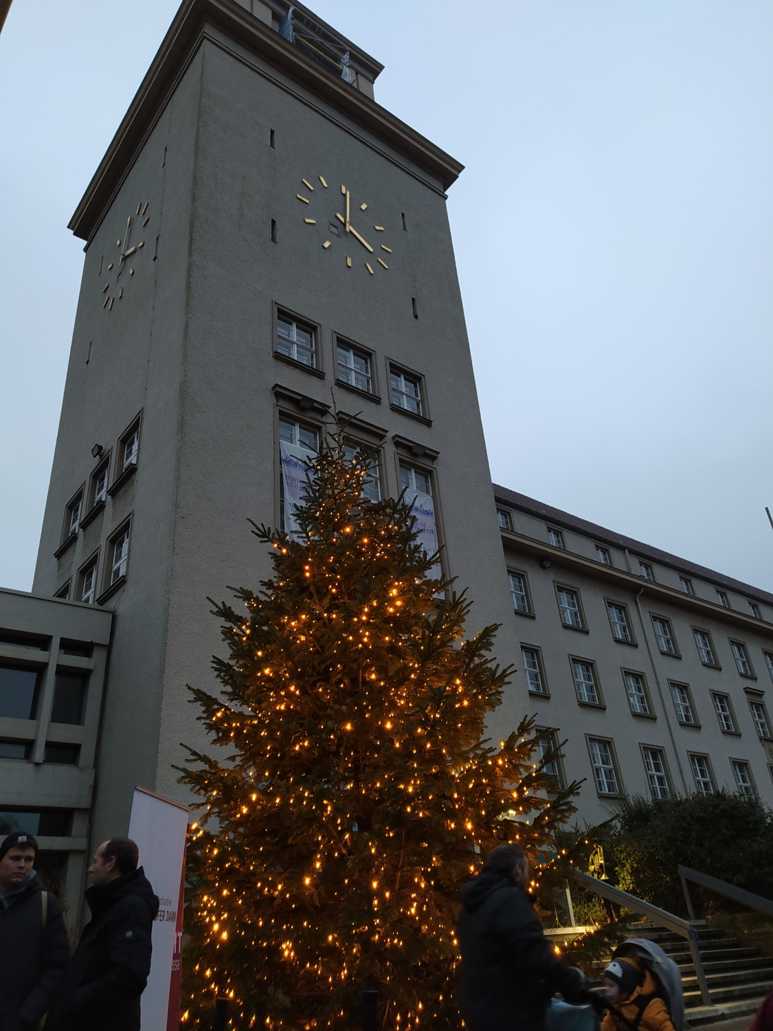 Foto im Hochformat mit dem Turm des Rathauses Tempelhof im Hintergrund, im Vordergrund ist ein beleuchteter Weihnachtsbaum, davor sieht man dunkel ein paar Menschen