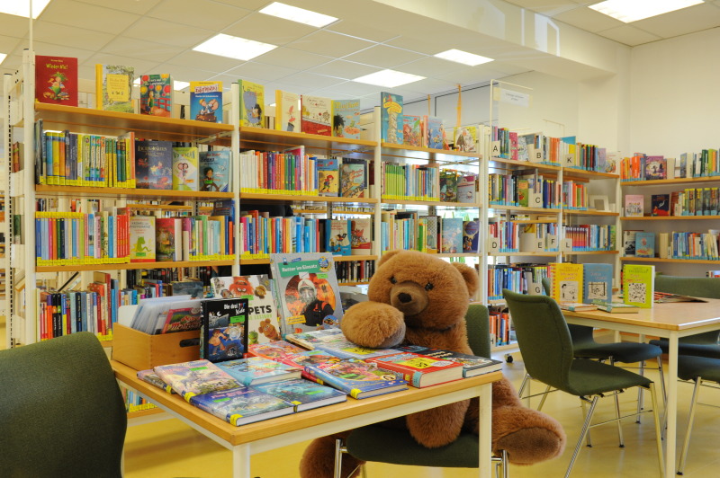 Blick auf einen teil des Kinderbereiches. Auf dem Tisch liegen Medien aus. Der Teddy sitzt daneben. Im Hintergrund stehen Regale mit Kinderbüchern.