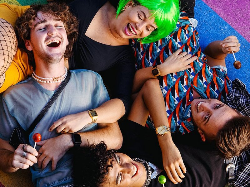 Eine Gruppe queere Jugendliche liegen auf einer gemalten Pride-Fahne.