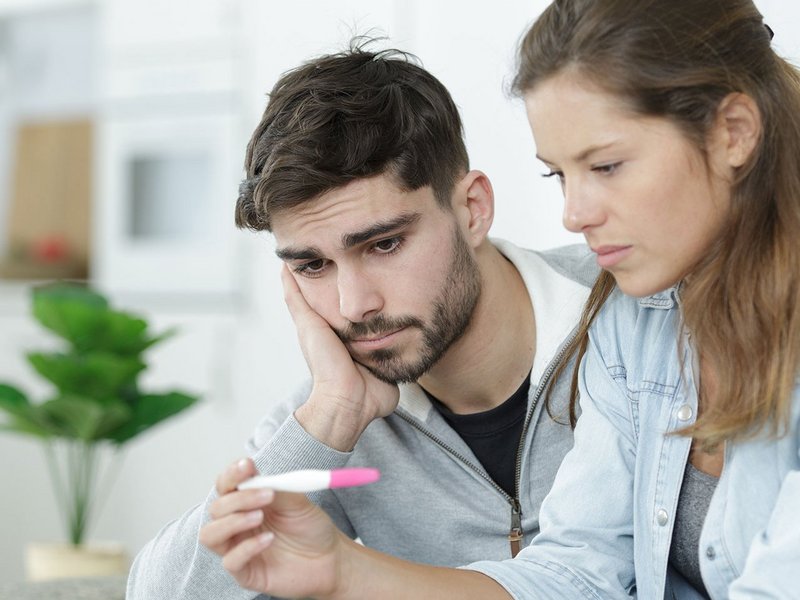Ein junges Paar sitzt nebeneinander und schaut traurig auf einen Schwangerschaftstest.