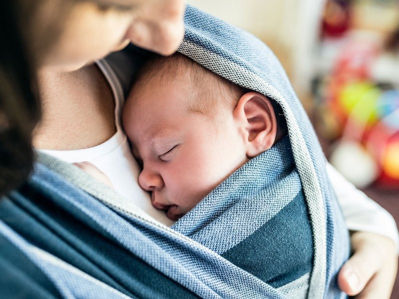 Eine Frau hält mit einem Tragetuch einen schlafenden Säugling auf dem Arm.