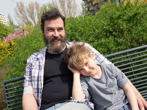 Ein Mann und ein Kind sitzen auf einer Parkbank vor grünen Büschen und einem Wohngebäude
