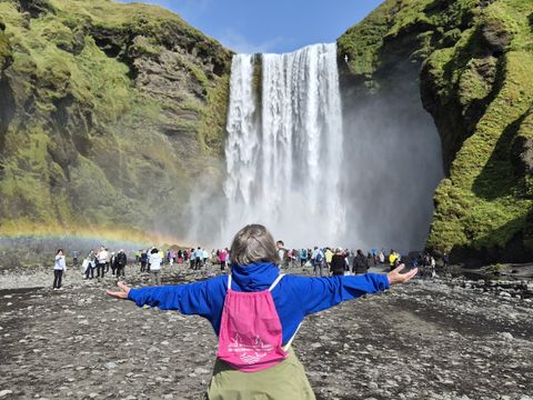 Im Vordergrund steht eine Person mit ausgebreiteten Armen, die einen pinken Rucksack trägt und auf einen imposanten Wasserfall blickt. Der Wasserfall stürzt von einer steilen, moosbedeckten Felswand herab. Zahlreiche Menschen stehen auf dem steinigen Boden vor dem Wasserfall, einige fotografieren oder betrachten das Naturschauspiel. Links im Bild ist ein Regenbogen zu sehen, der sich durch die feuchte Luft bildet. Die Szene spielt auf Island und vermittelt die beeindruckende Naturkulisse mit grünen Felsen und klarem Himmel.