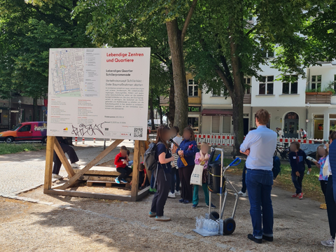 Baustellenschild vom Lebendigen Quartier Schillerpromenade: Frau spricht in Mikrofon und Kinder hören zu, verschwommene Gesichter