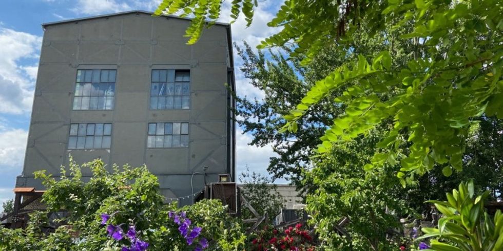 Im Vordergrund blühen verschiedene Pflanzen und Bäume in einem Garten. Dahinter steht ein Industriebau mit vielen Fenstern.