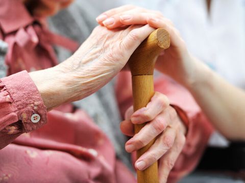 Eine alte Frau hält sitzend einen Gehstock mit der linken Hand umfasst und hat die rechte Hand auf den Griff gelegt. Darüber ist die Hand einer jüngeren Pflegeperson zu sehen.