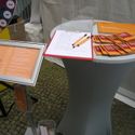 Bildvergrößerung: Ein Standtisch mit den Minibooklets, dem Gästebuch, Blätter und Stifte. Ein Teil vom Glücksrad und die Anleitung dazu.