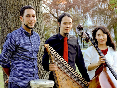 Drei Menschen stehen mit ihren Instrumenten in einem Park und lächeln in die Kamera.