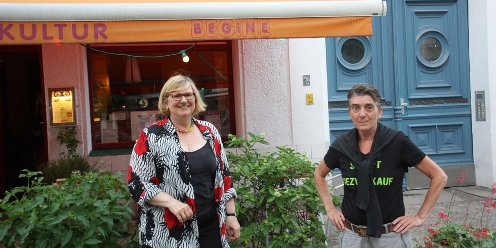 Zwei Personen stehen vor einem Café. Auf dem Sonnenschutz des Cafès steht Kultur und Begine.