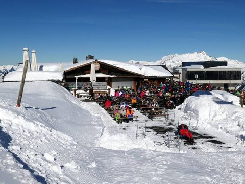 verschneite Berglandschaft mit Baude im Vordergrund davor sitzen Personen welche essen und trinken