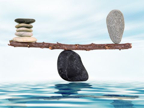 Auf einem Stein liegt ein Ast. Auf der linken Seite liegen mehrere flache Steine übereinander auf dem Ast. Auf der rechten Seite hält ein größerer Stein das Gleichgewicht.