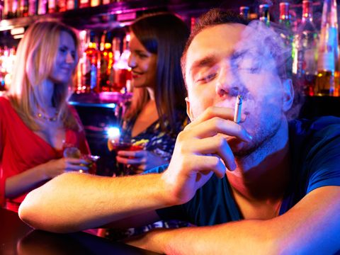 Mann raucht Zigarette in Bar, zwei junge Frauen unterhalten sich im Hintergrund
