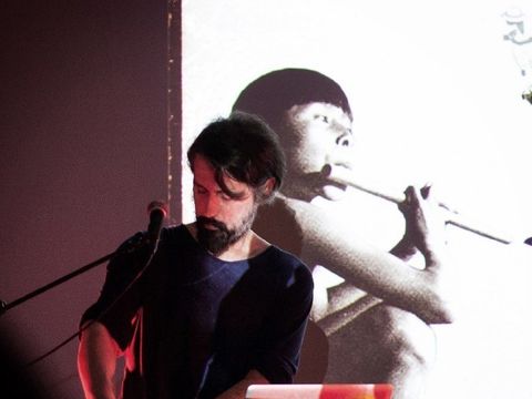 Bildvergrößerung: Ein Mann steht an einem DJ-Pult. Hinter ihm hängt ein schwarz-weißes Foto auf dem eine Junge Person Querflöte spielt.