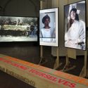 Bildvergrößerung: Blick in die Ausstellung mit einer Holzbank mit Texten darauf, Monitoren mit Porträts von Frauen und im Hintergrund eine breite Projektionsleinwand, auf der Frauen aus der Zeit um 1918 Bretter mit Schriftzügen hochhalten