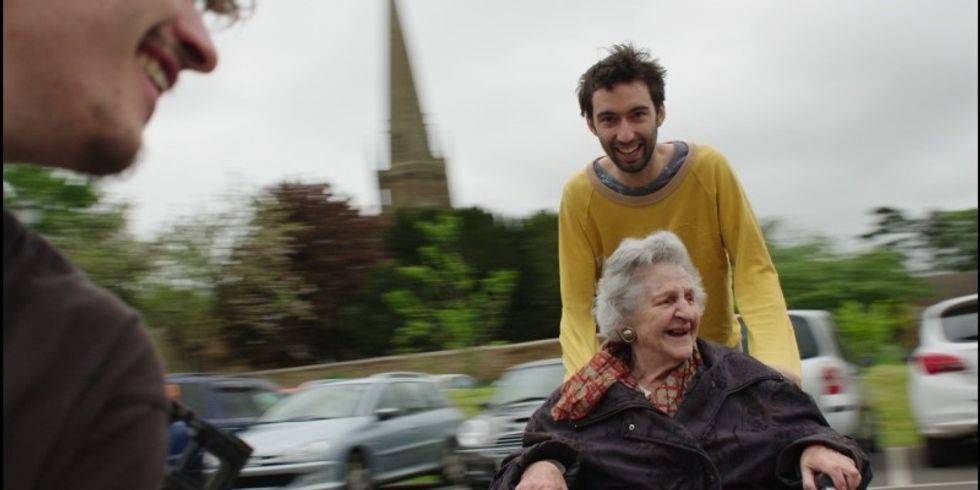 Ein leicht verschwommenes Foto, das eine ältere Dame im Rollstuhl zeigt, die von einem jungen Mann geschoben wird. Beide lächeln.