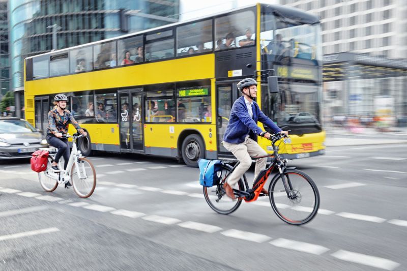 Zwei E-Bikes überqueren die Straße. Dahinter fährt ein Bus. Im linken Rand des Bildes sieht man die Kühlerhaube eines fahrenden Autos