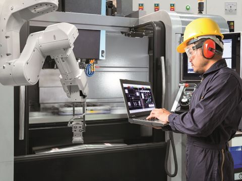 Ein Mann in Schutzkleidung steht mit einem Laptop in der Hand vor einem Roboterarm