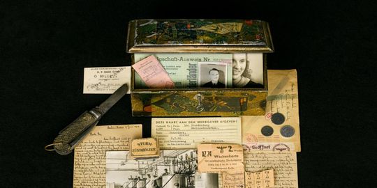 Auf dem Foto ist eine geöffnete Blechdose zu sehen. In der Dose befinden sich alte Fotos und Dokumente. Neben der Dose liegen ebenfalls alte Fotos und Dokumente aus der Nazi-Zeit sowie ein geöffnetes Taschenmesser und drei Münzen.