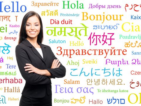 Frau im schwarzen Blazer steht lächelnd vor Wörtern (unterschiedliche Sprachen für "Hallo")