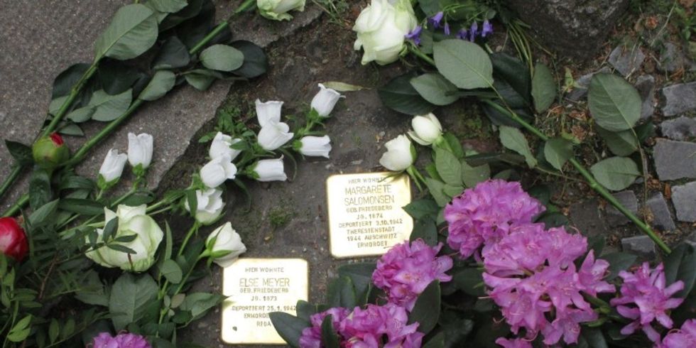 Ein Verlege-Beispiel von Stolpersteinen: Zwei Stolpersteine von oben fotografiert, daneben liegen weiße und lilane Blumen.