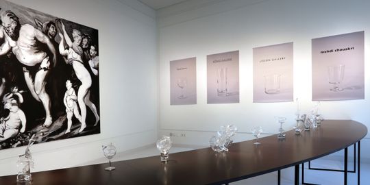 Vorn sind aus Glasbruchstücken neu zusammengesetzte Objekte auf einem schwarzen halbrunden Tisch zu sehen. Dahinter links hängt ein großformatiges schwarz-weißes Gemälde an der Wand. Rechts an der Wand hängen 4 Prints auf denen jeweils ein Glas und ein bekannter Kunstgalerienname abgebildet ist. 