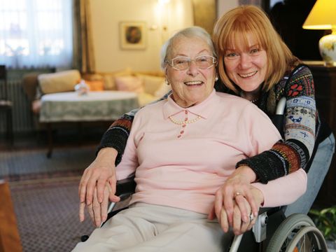 Seniorin im Rollstuhl wird von einer jüngeren Frau umarmt. Beide lächeln in die Kamera.