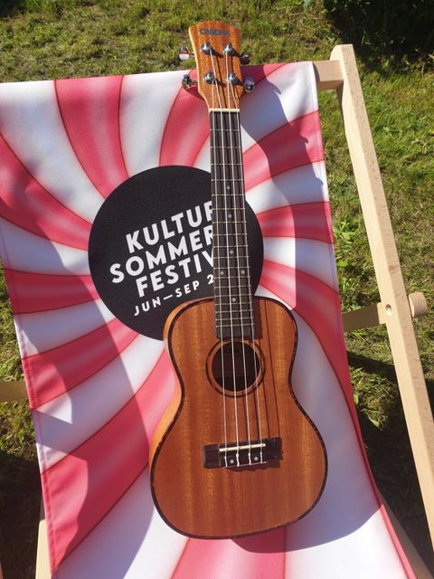 Bildvergrößerung: Ein rot-weiß gestreifter Liegestuhl mit Logo des Kultursommerfestivals 2023 auf der eine Ukulele liegt.