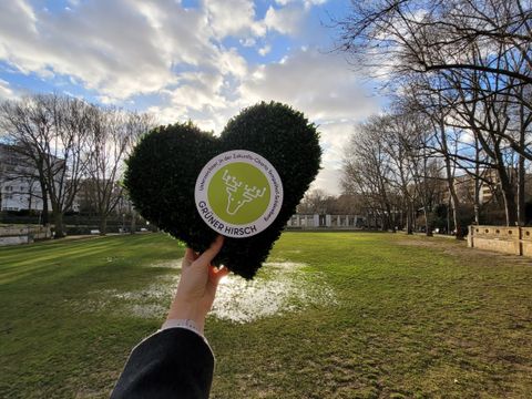 Eine Hand hält ein grünes Herz mit dem Logo des Grünen Hirschen in die Luft. Im Hintergrund ist ein Park zu sehen. 
