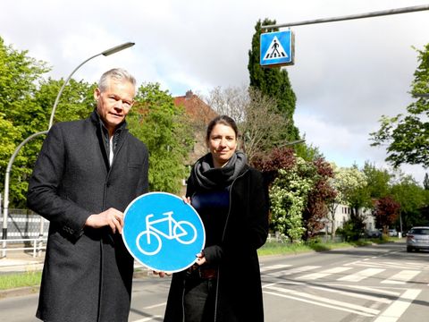 Bildvergrößerung: Ein Mann und eine Frau stehen an einem Straßenrand und halten ein Schild für einen Fahrradweg in den Händen. Hinter den beiden ist ein Zebrastreifen.