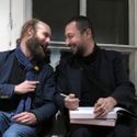 Bildvergrößerung: Nihad Nino Pusija mit seinem Galeristen im Gespräch, die zwei sitzen vor dem Fenster und lachen herzlich, während Pusija seine Publikation in den Händen hält und im Begriff ist, diese zu signieren.