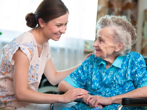 Junge Pflegerin sitzt lächelnd einer Seniorin gegenüber und streicht über deren Schulter