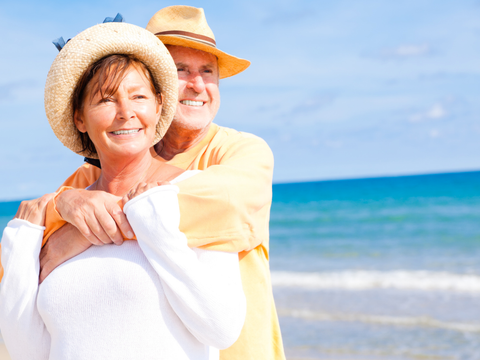 eine ältere Frau und ein älterer Mann im sommerlichen Outfit umarmen sich am Meer