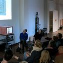 Bildvergrößerung: Ein Sprecher erläutert ein auf die Wand projiziertes Bild für ein sitzendes Publikum