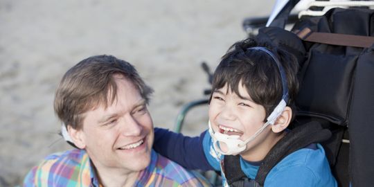 Ein Vater sitzt lächelnd neben seinem behinderten Sohn am Strand