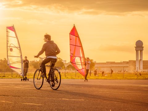 In der Bildmitte ein Mann auf einem Fahrrad. Rechts und links davon zwei Windsurfer, die auf der Startbahn des Tempelhofer Feld unterwegs sind. Im Hintergrund der ehemalige Tower des Flughafens