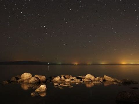 Fluss bei Nacht Felsen scheinen im Vordergrund durch die Wasseroberfläche am Horizont strahlen verschiedene Lichtquellen je weiter nachoben von den Lichtquellen entfernt nimmt die Zahl sichtbarer Sterne zu