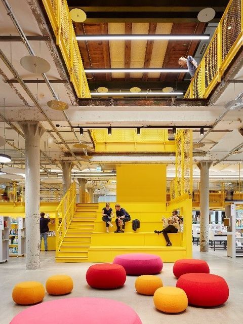 Bildvergrößerung: In einer Halle sitzen auf einer gelben Treppe 3 Menschen und lesen. Im Vordergrund sind mehrere unterschiedlich große Sitzkissen.