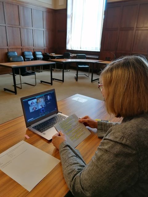 Bildvergrößerung: Frau Schöttler sitzt an einem Tisch vor einem Laptop, auf dem einige Ateur_inne zu sehen sind und hält Karteikarten mit ihrem Text in den Händen.