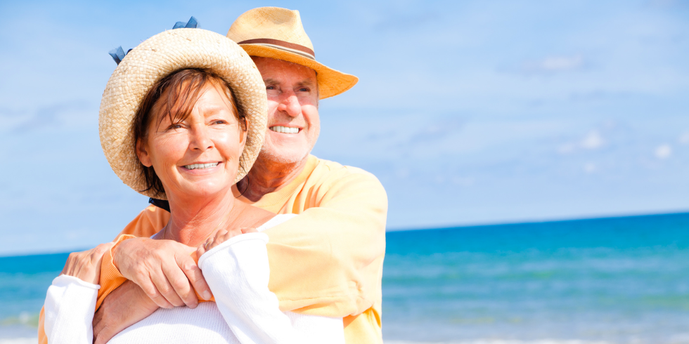 eine ältere Frau und ein älterer Mann im sommerlichen Outfit umarmen sich am Meer