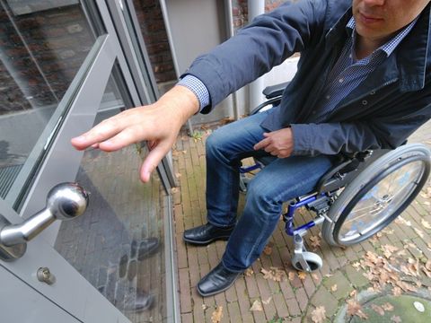 Ein Mann in einem Rollstuhl greift nach der Türklinke einer geschlossenen Tür