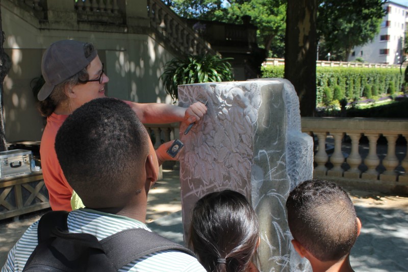 Die Bildhauerin Roswitha Schaab arbeitet an ihrem Stein im Körnerpark, drei Kinder sehen ihr dabei zu.