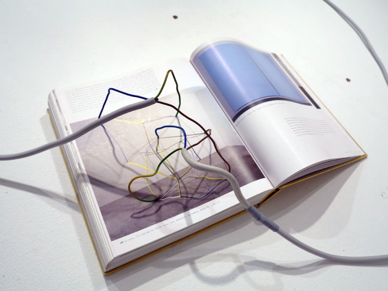 aufgeschlagenes Buch mit der Abbildung einer Skulptur, darauf befinden sich Elektrokabel, die wie eine Verlängerung der Skulptur wirken