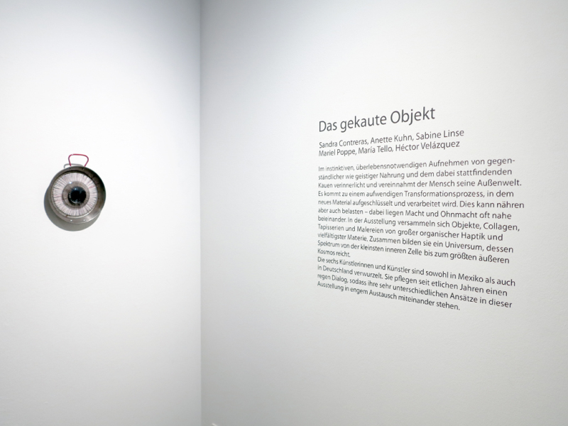 Ansicht einer Raumecke, auf der linken Wand befindet sich ein augenförmiges Objekt aus einer Paellapfanne, Winkelmessern und Nähseide, auf der rechten Wand befindet sich der in Dunkelgrau geplottete Ausstellungstext zur Ausstellung "Das gekaute Objekt".