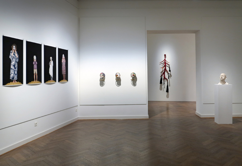 Eine Ansicht des Galkerieraumes, links eine Reihe von fotografien an der Wand, daneben drei totenkopfartige Skulpturen, im Hintergrund eine gehäkelte Skulptur mit schwarz-roten Tentakeln, rechts davon eine Männerbüste aus Gips auf einem weißen Sockel.