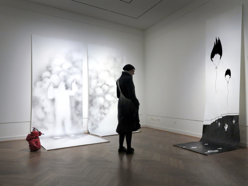 Eine Besucherin betrachtet die beiden großformatigen Arbeiten. Inside zeigt eine auf zwei Papierbahnen aufsteigende Wolke mit der weißen Silhouette einer Person. Daneben steht eine surreale Figur, die aus einem Feuerlöscher heraus entstanden ist.