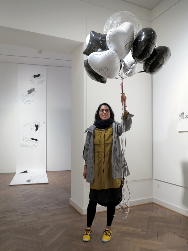 Die Künstlerin hält aufsteigende schwarze und weiße Luftballons in der Hand. Darauf erkennt man gezeichnete Gesichter .