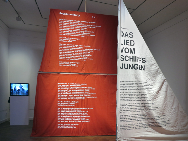 Im Vordergrund ein weißes Segel mit dem Text "Das Lied vom Schiffsjungen" von Margarete Steffin, dahinter ein rotes Segel mit dem Text "Seeräuber-Jenny" von Bertolt Brecht.